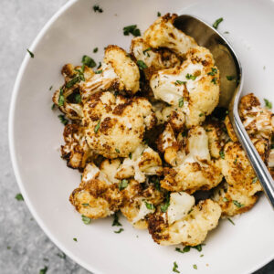 Air fryer roasted cauliflower in a white bowl with a silver spoon, garnished with fresh herbs.