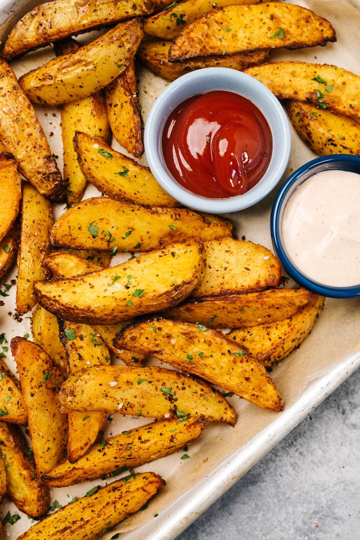 Air fryer potato wedges on a parchment lined baking sheet with small dip bowls of ketchup and fry sauce.