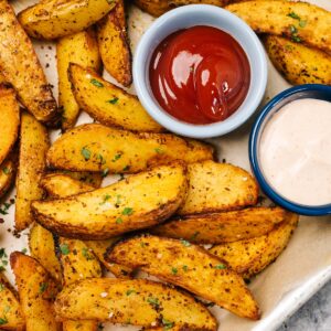 Air fryer potato wedges on a parchment lined baking sheet with small dip bowls of ketchup and fry sauce.