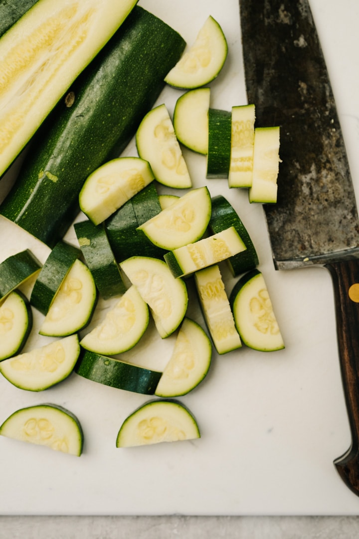 Raw zucchini chopped into half moon shapes on a white cutting board with a large chef's knife.