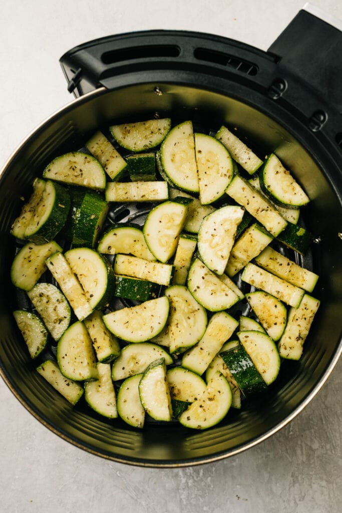 Chopped zucchini tossed with olive oil, garlic, and italian seasoning in the basket of an air fryer.