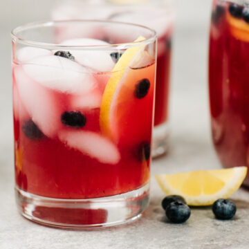 Two glasses and a pitcher of blueberry lemonade on a concrete background with lemon wedges and fresh blueberries scattered around the glasses.