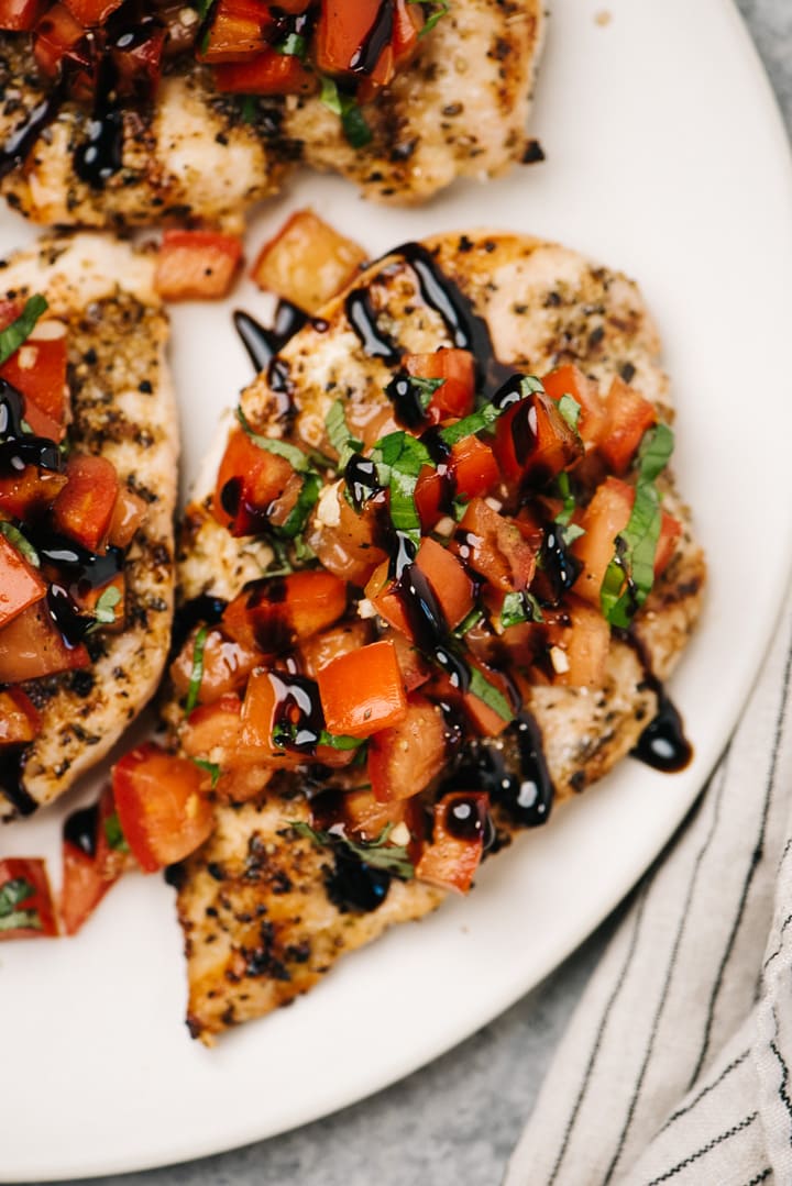 Three grilled chicken breasts topped with tomato bruschetta and garnished with balsamic glaze on a cream serving platter with a striped linen napkin.