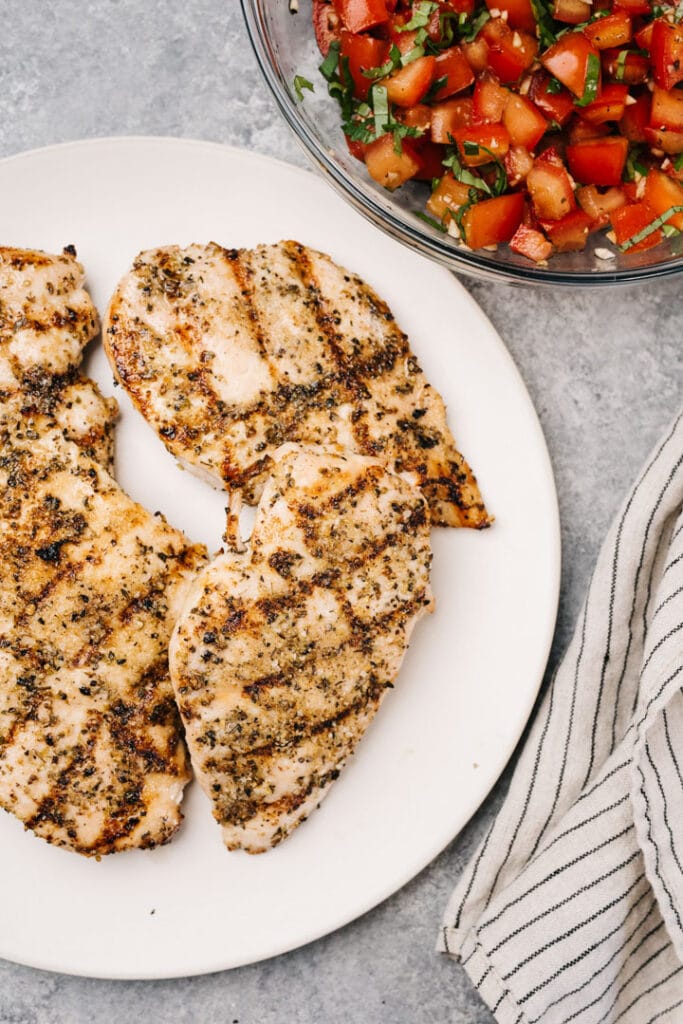 Four grilled chicken breasts on a cream serving plate with a bowl of tomato bruschetta and a striped linen napkin to the side.