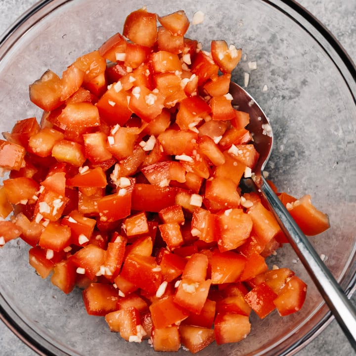 Chopped tomatoes mixed with minced garlic and salt in a glass mixing bowl with a spoon.