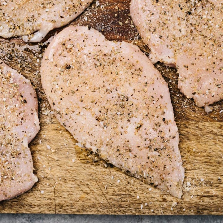 Four boneless skinless chicken breasts brushed with olive oil and seasoned with salt, pepper, and herbs on a wood cutting board.