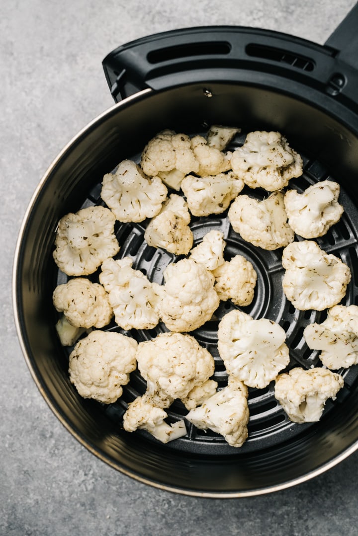 Raw cauliflower florets tossed with olive oil, salt, and pepper in the basket of an air fryer on a concrete background.