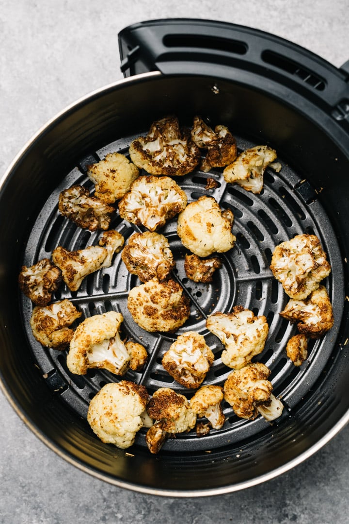 Crispy roasted air fried cauliflower florets in the basket of an air fryer on a cement background.