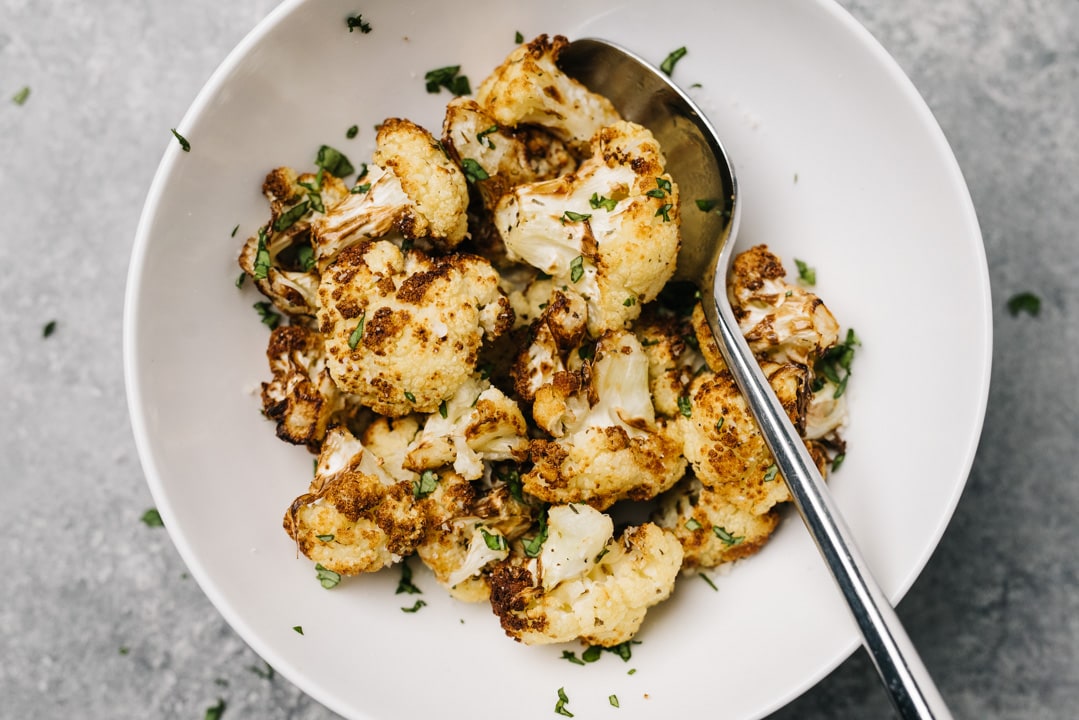 Air fryer cauliflower in a white bowl with a silver spoon, garnished with fresh herbs.