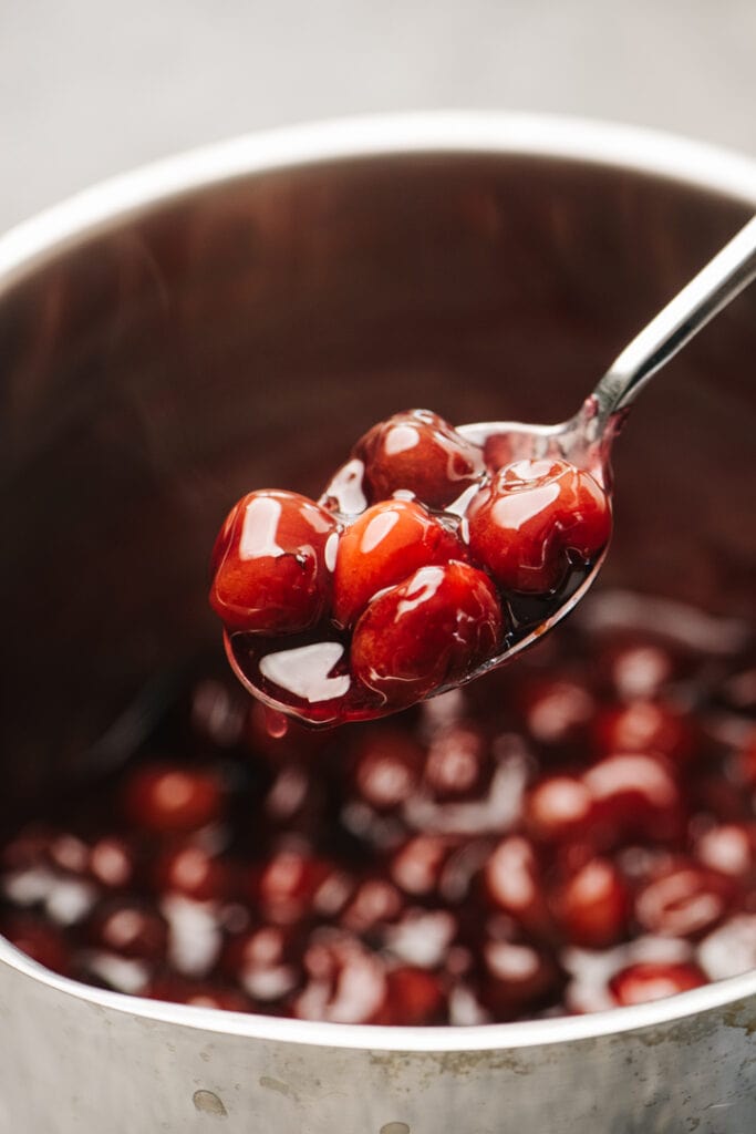 A spoonful of homemade cherry pie filling hovering over a sauce pot.
