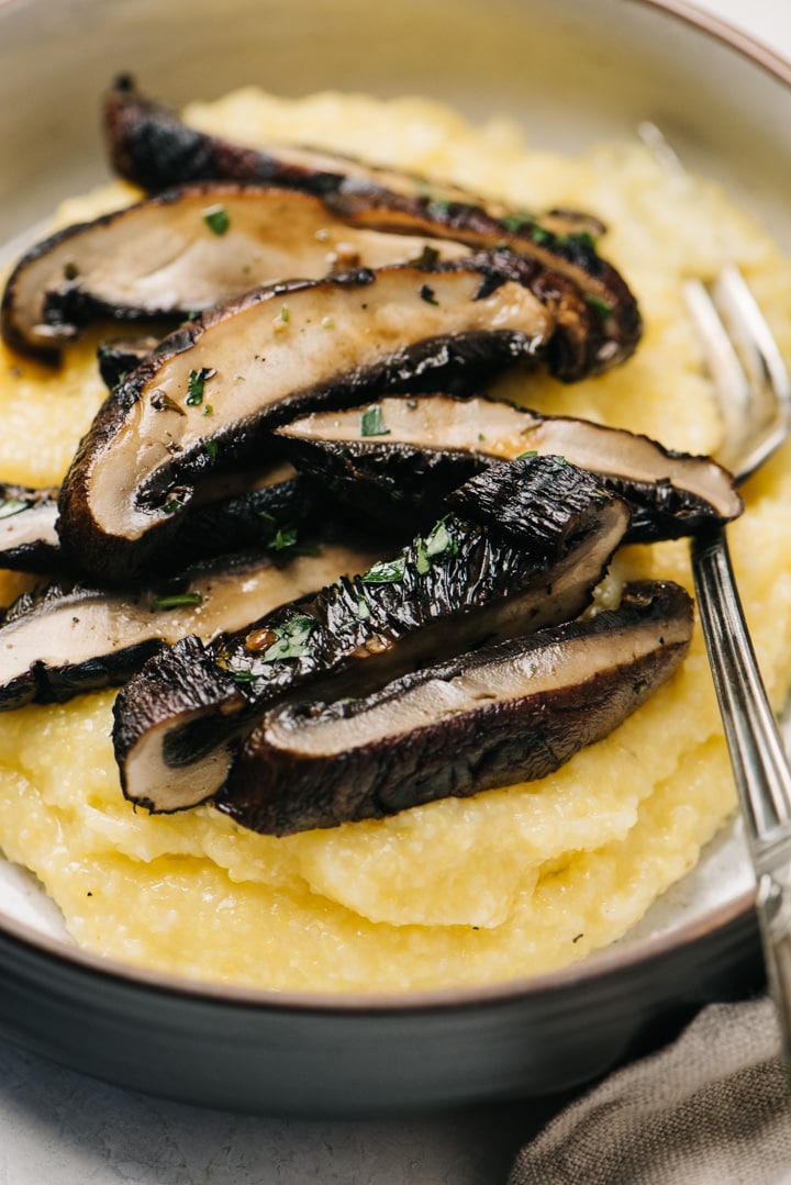 Side view, sliced portobello mushrooms over polenta in a tan and black bowl with silver fork.