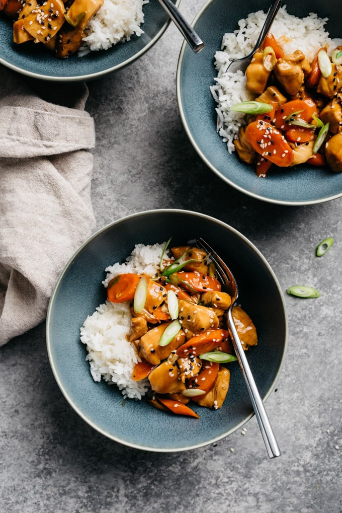 Three blue bowls of honey garlic chicken over white rice on a cement background with a tan linen napkin.