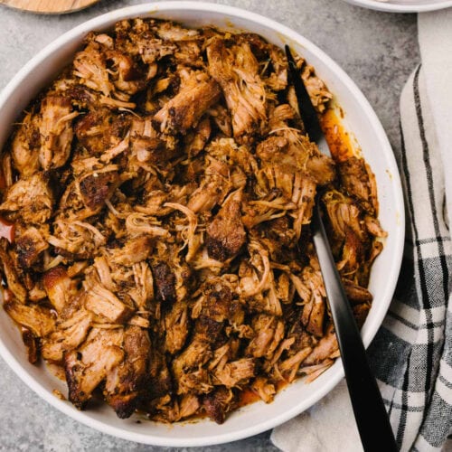 A black serving fork tucked into a white serving bowl of pulled pork with a linen napkin to the side.