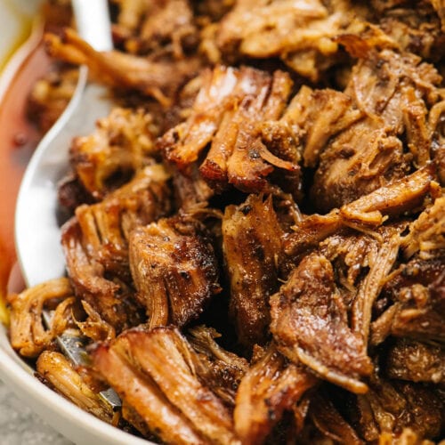 Side view, shredded pulled pork in a tan serving bowl with a silver serving fork.