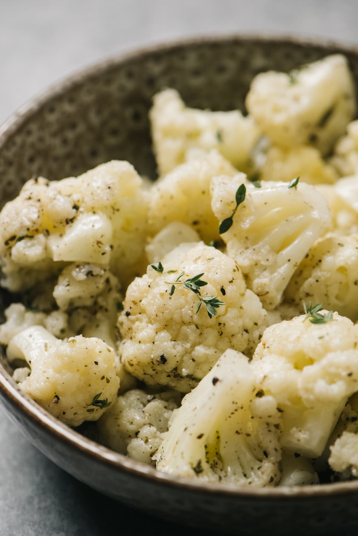Side view, instant pot cauliflower florets tossed with fresh herbs in a brown speckled bowl on a concrete background.