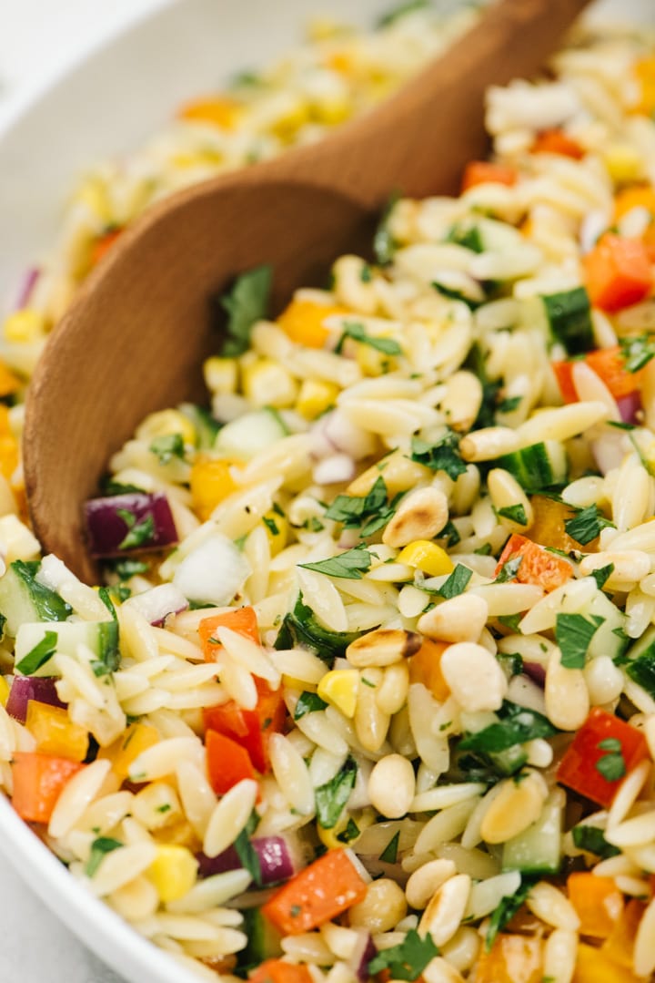 Side view, a wood serving spoon tucked into a bowl of mediterranean orzo salad.