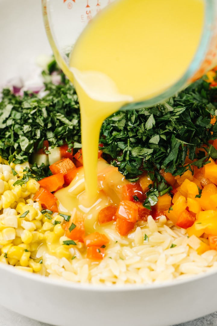 Pouring lemon vinaigrette over orzo salad with finely chopped herbs and vegetables.