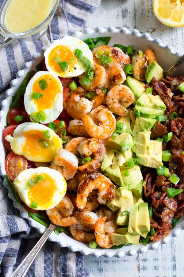 Spring cobb salad with runny eggs, shrimp, avocado, and bacon in a white serving bowl with a blue striped napkin.