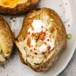 Three air fryer baked potatoes on a white plate with various toppings, including sour cream, bacon bits, shredded cheese, and chives.