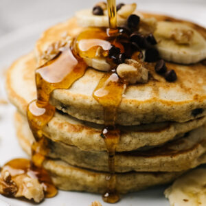 Pouring maple syrup onto a stack of banana bread pancakes, topped with sliced bananas, walnuts, and mini chocolate chips.