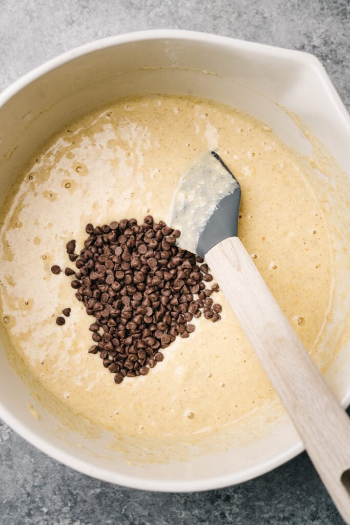 Mini chocolate chips added to banana bread pancake batter in a large mixing bowl with a rubber spatula.