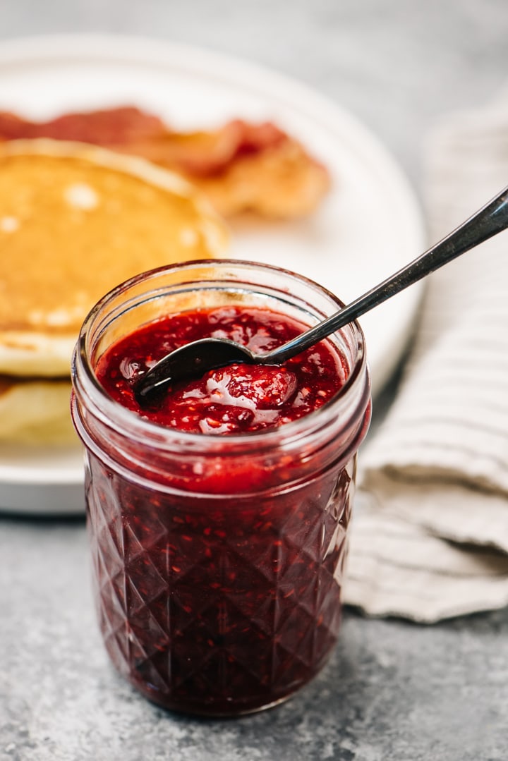 Side view, a jar of berry compote with a plate of pancakes in the background.