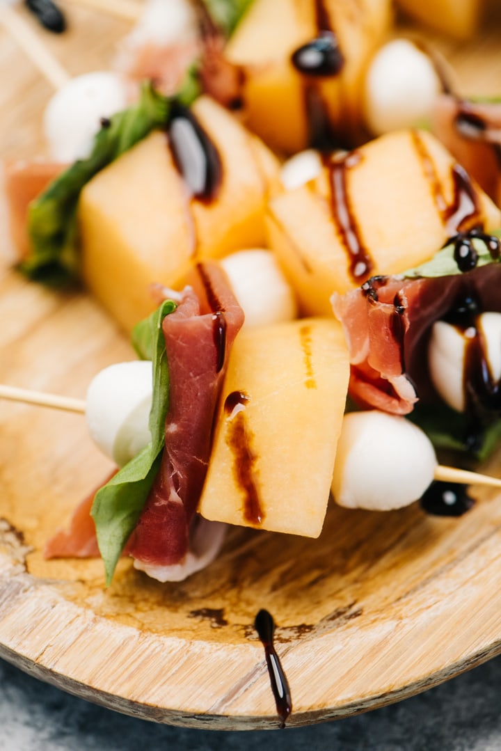 Side view, cantaloupe and prosciutto appetizer bites on a wood platter, drizzled with balsamic glaze.