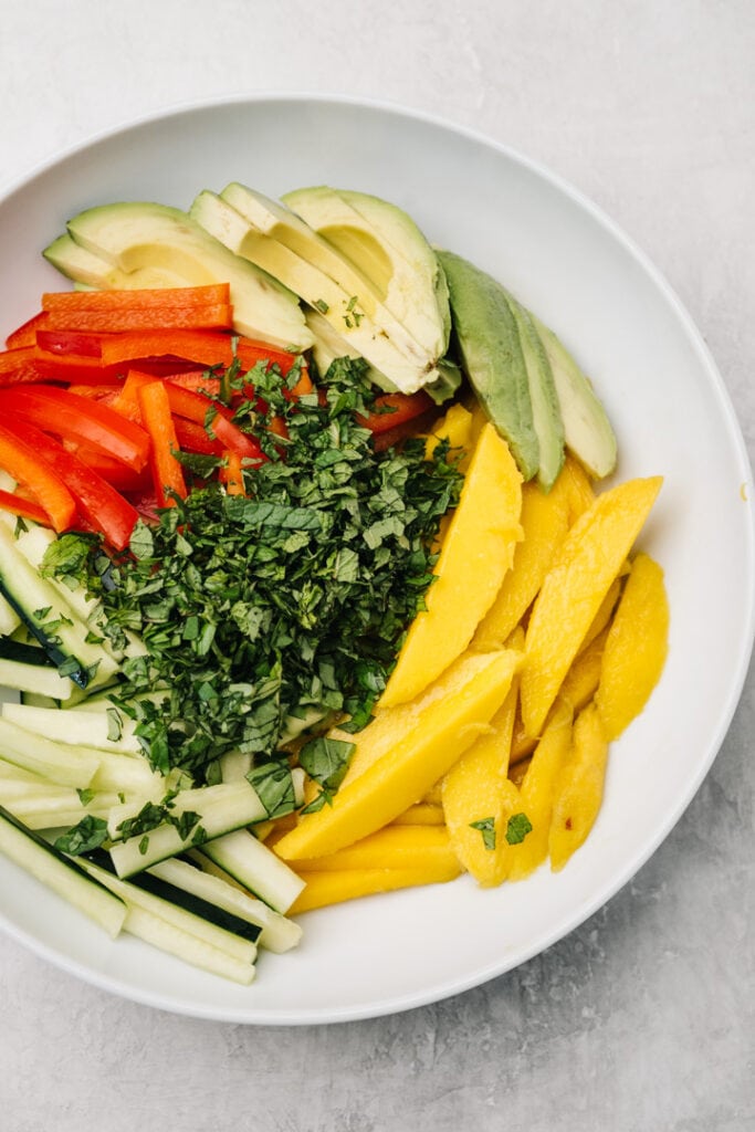 Sliced mangos, avocado, bell pepper, and cucumber in a white mixing bowl topped with minced basil and mint.