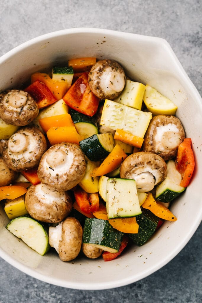 Mushrooms, squash, zucchini, and bell peppers tossed with marinade in a large mixing bowl.