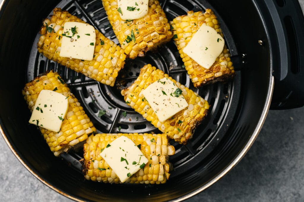 Air fryer corn in an air fryer basket topped with butter, salt, and pepper.