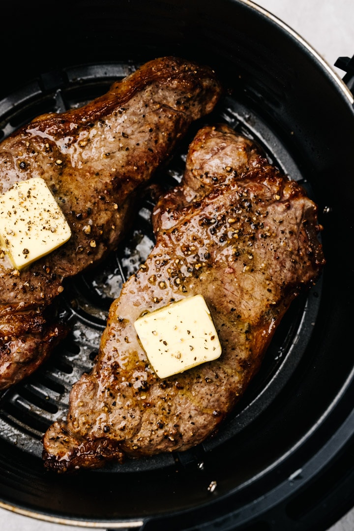Two cooked steaks in the basket of an air fryer topped with small pats of butter.