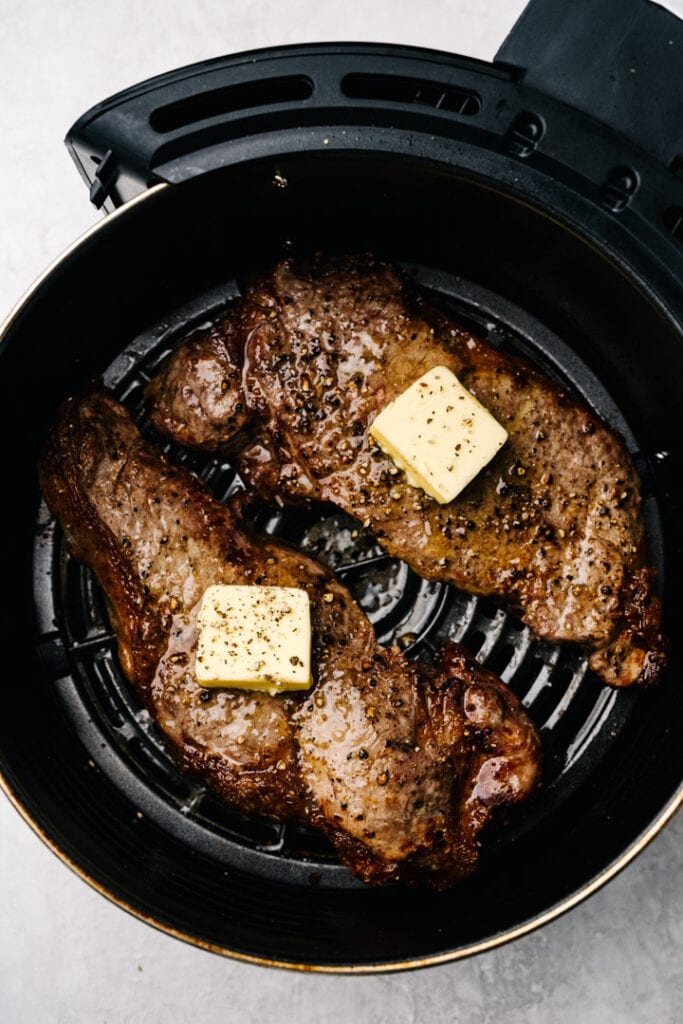 Two cooked steaks in the basket of an air fryer topped with small pats of butter.