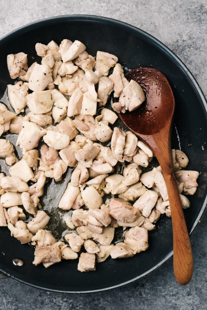 Diced chicken thigh sautéing in a skillet with a wood spoon.