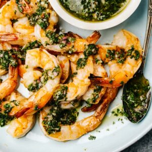 Grilled shrimp drizzled with chimichurri sauce on a blue serving plate with a small bowl of sauce to the side.