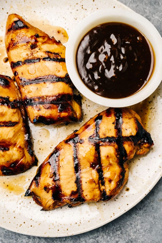 Three grilled teriyaki chicken breasts on a platter with a small bowl of teriyaki sauce.