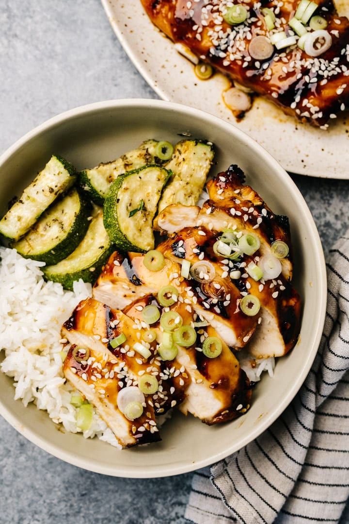 Grilled teriyaki chicken slices over white rice in a tan bowl with a platter of chicken in the background.