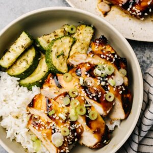 Slices of grilled teriyaki chicken over white rice and sautéed zucchini, garnished with green onions and sesame seeds.