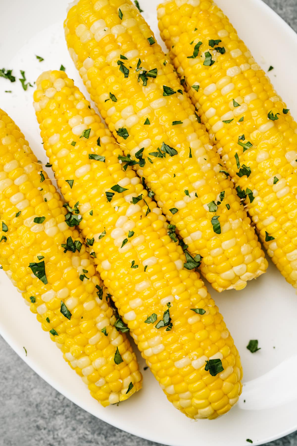 Four ears of cooked corn on the cob seasoned with butter and fresh herbs on a white plate.
