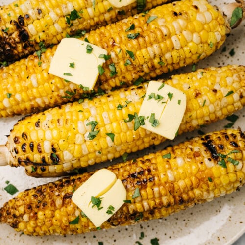 Four ears of cooked corn on the cob seasoned with butter and fresh herbs on a tan speckled plate.