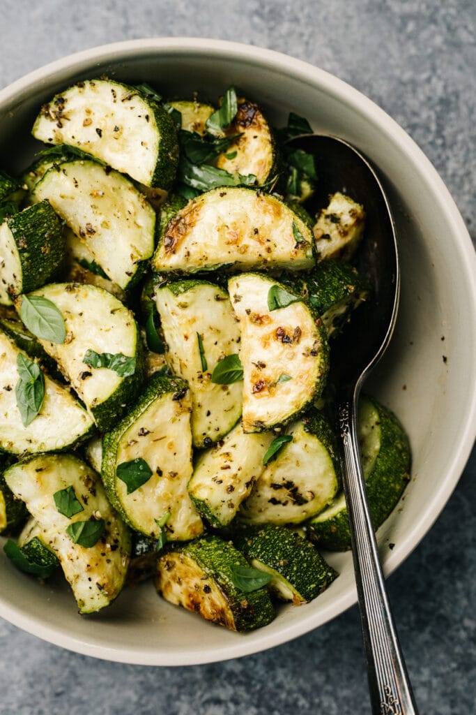 Roasted zucchini in a tan serving bowl with a silver serving spoon.