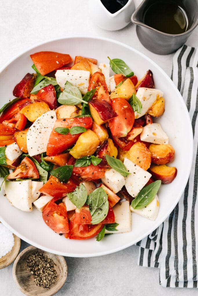A peach caprese salad in a white salad bowl with olive oil and balsamic on the side next to a striped linen napkin.
