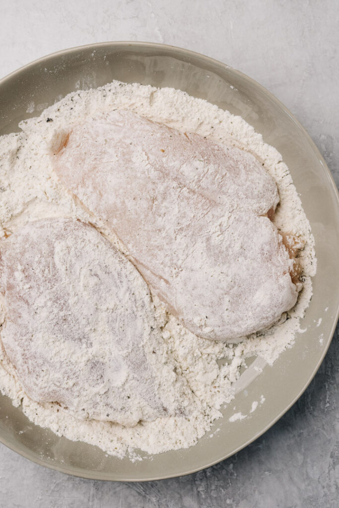 Two chicken breasts coated with flour and seasonings in a tan bowl.