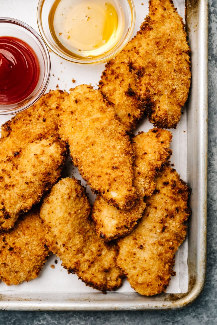 Homemade chicken tenders made in the airy fryer on a baking sheet with small dip bowls of ketchup and honey.