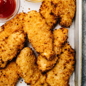 Air Fryer chicken tenders with buttermilk arrange on a baking sheet with ketchup on honey in glass dipping bowls.