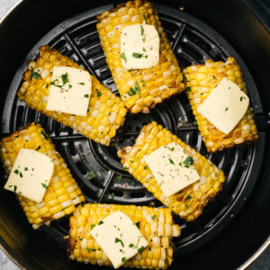 Air fryer corn on the cob in the basket of an air fryer, topped with butter and chopped basil.