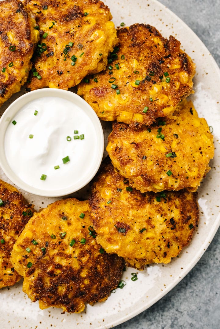 Corn fritters with bacon arranged on a speckled tan plate with a small bowl of sour cream for dipping.