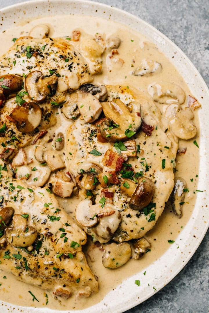 Chicken breasts with mushrooms and bacon in a creamy sauce on a tan speckled plate.
