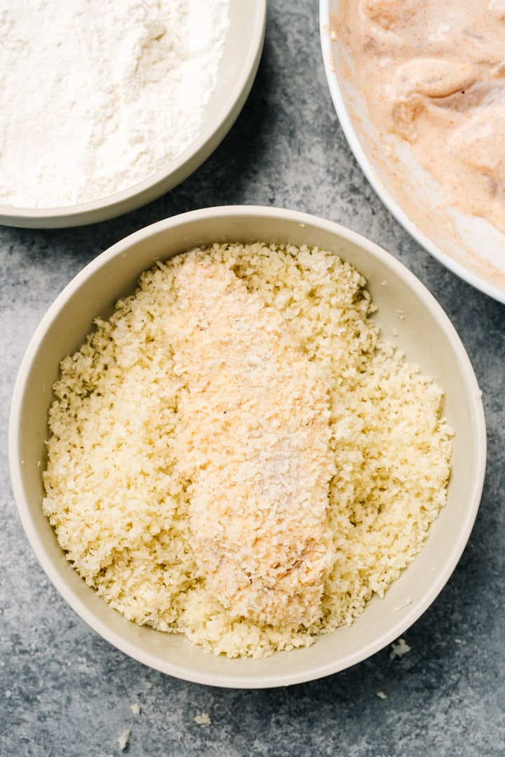 A chicken tenderloin being dredged in panko breadcrumbs in a shallow bowl.
