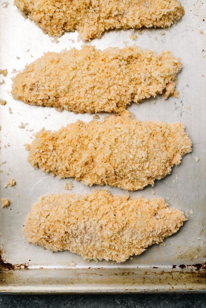 Chicken tenderloins coated with flour and breadcrumbs on a baking sheet.