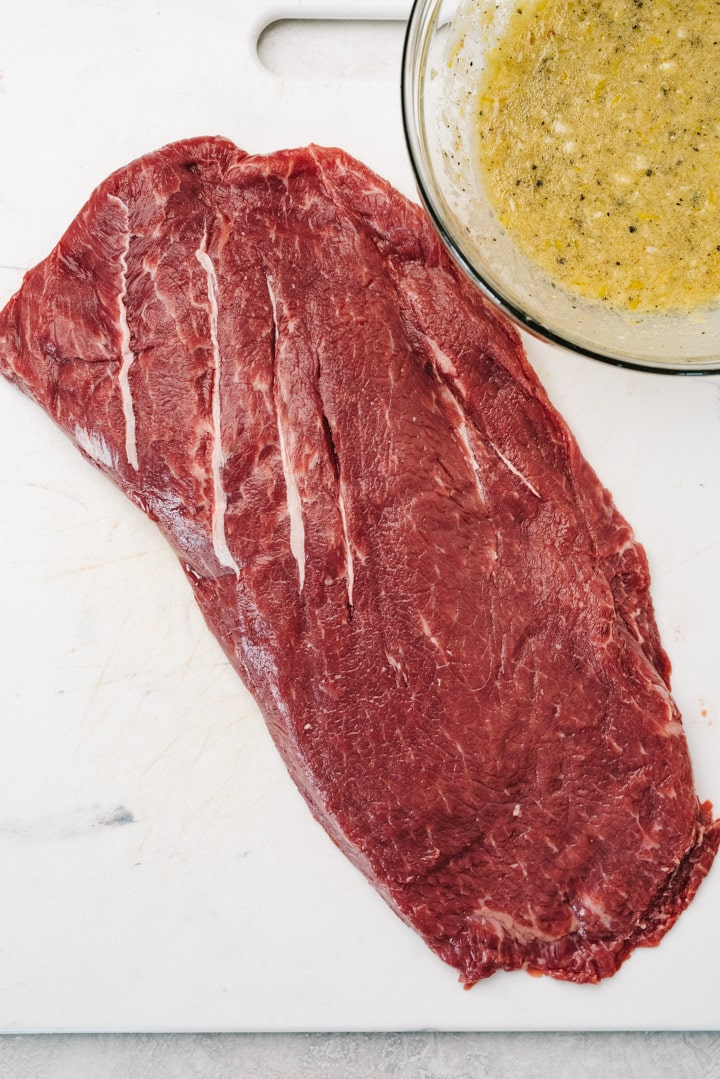 A raw flat iron steak on a cutting board next to a small bowl of marinade.